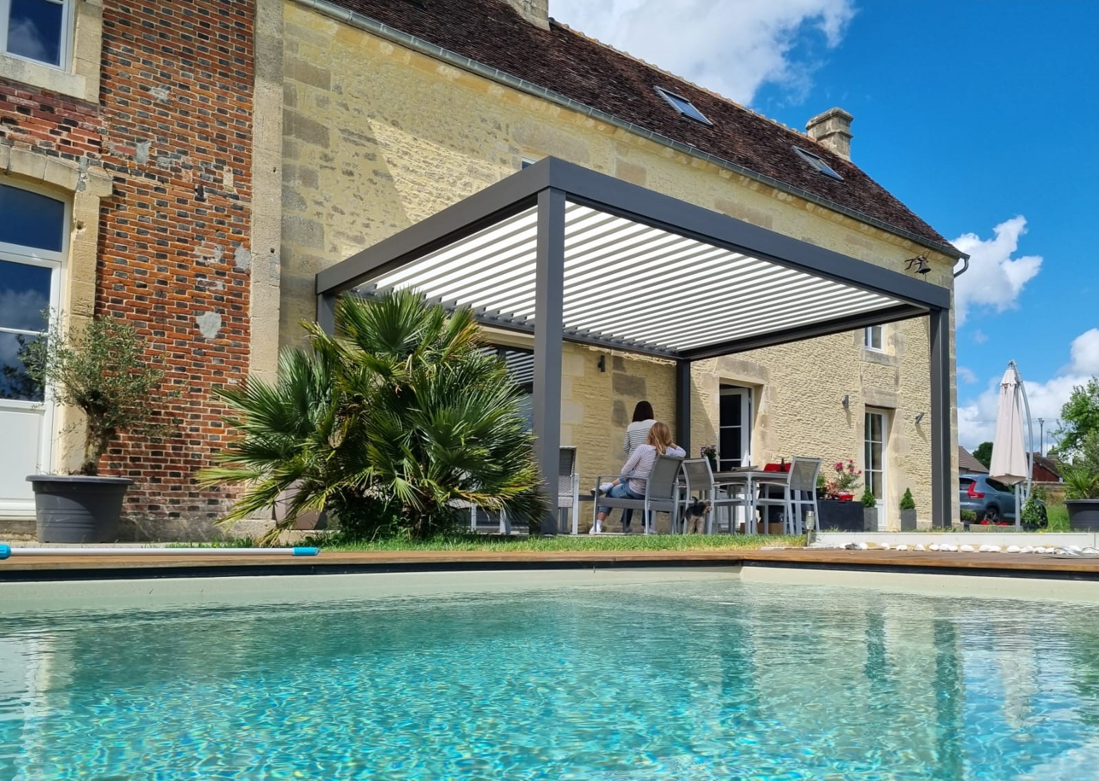 pergola bioclimatique posée sur une maison traditionelle en normandie avec une famille installée en-dessous sur un salon de jardin au bord d'une piscine
