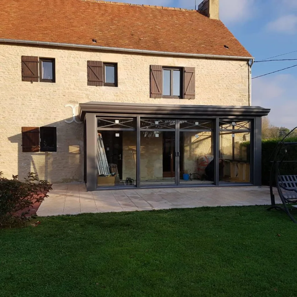 véranda aluminium normandie concept alu 
couleur gris clair poosée sur une maison ancienne normande 